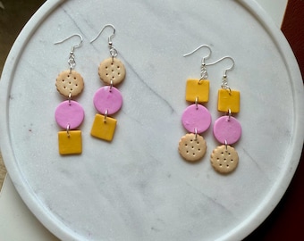 May include: Two pairs of dangle earrings with a playful design. Each earring features a combination of shapes: a beige, round cracker, a pink circle, and a yellow square. The earrings are displayed on a white marble tray.