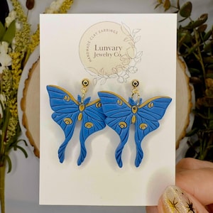 May include: A pair of blue and gold butterfly earrings made of polymer clay. The earrings are handmade and feature a gold hook closure. The earrings are labeled "Lunvary Jewelry Co."