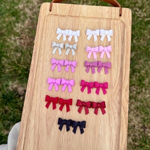 May include: A wooden tray with a variety of small, handmade bows in different colors, including white, pink, red, and black. The bows are arranged in rows on the tray, with the lightest colors at the top and the darkest colors at the bottom.
