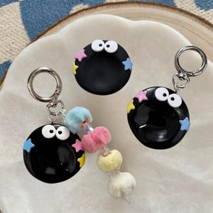 May include: Three black round keychains with white eyes and colorful star accents. One keychain has a fluffy, multi-colored charm. The keychains have silver-colored metal clasps.