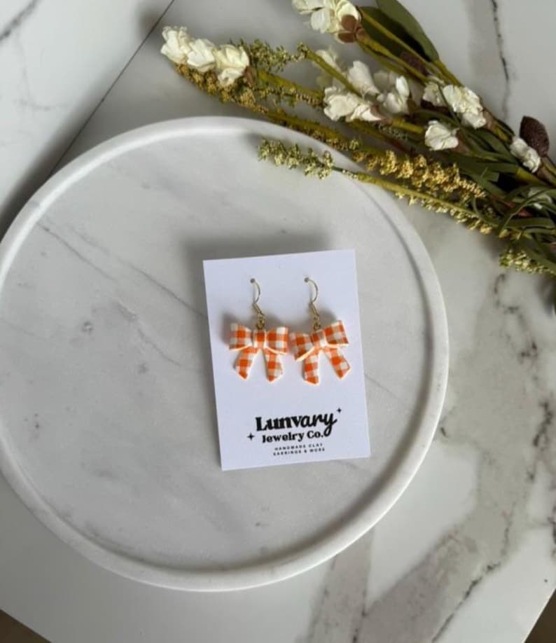 May include: A pair of orange and white gingham bow earrings with gold-colored hooks. The earrings are displayed on a white card with the text "Lunvary + Jewelry Co.". The earrings are on a white marble tray with dried flowers.