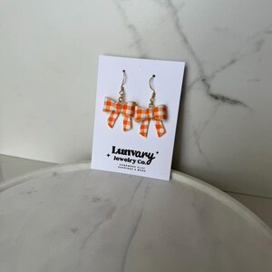 May include: A pair of bow-shaped earrings with a gold-colored hook. The bows are orange and white gingham. The earrings are displayed on a white card with the text "Lumvary + Jewelry Co. HANDMADE CLAY EARRINGS & MORE."