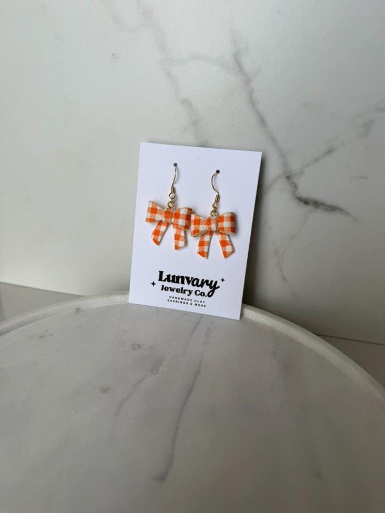 May include: A pair of bow-shaped earrings with a gold-colored hook. The bows are orange and white gingham. The earrings are displayed on a white card with the text "Lumvary + Jewelry Co. HANDMADE CLAY EARRINGS & MORE."