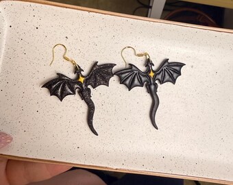 May include: A pair of black dragon earrings with gold-colored ear wires. The dragon has outstretched wings and a long, curved tail. The dragon's body has a yellow star-shaped accent.