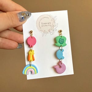 May include: A pair of handmade polymer clay earrings. One earring features a pink circle, a yellow and orange figure, and a rainbow. The other earring features a green hat with a shamrock, a blue crescent moon, and a pink figure.