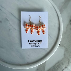 May include: A pair of bow-shaped earrings with a white and orange gingham pattern. The earrings are displayed on a white card with the text "Luvary Jewelry Co." and the words "HANDMADE CLAY EARRINGS & MORE". The earrings have gold-colored hooks.