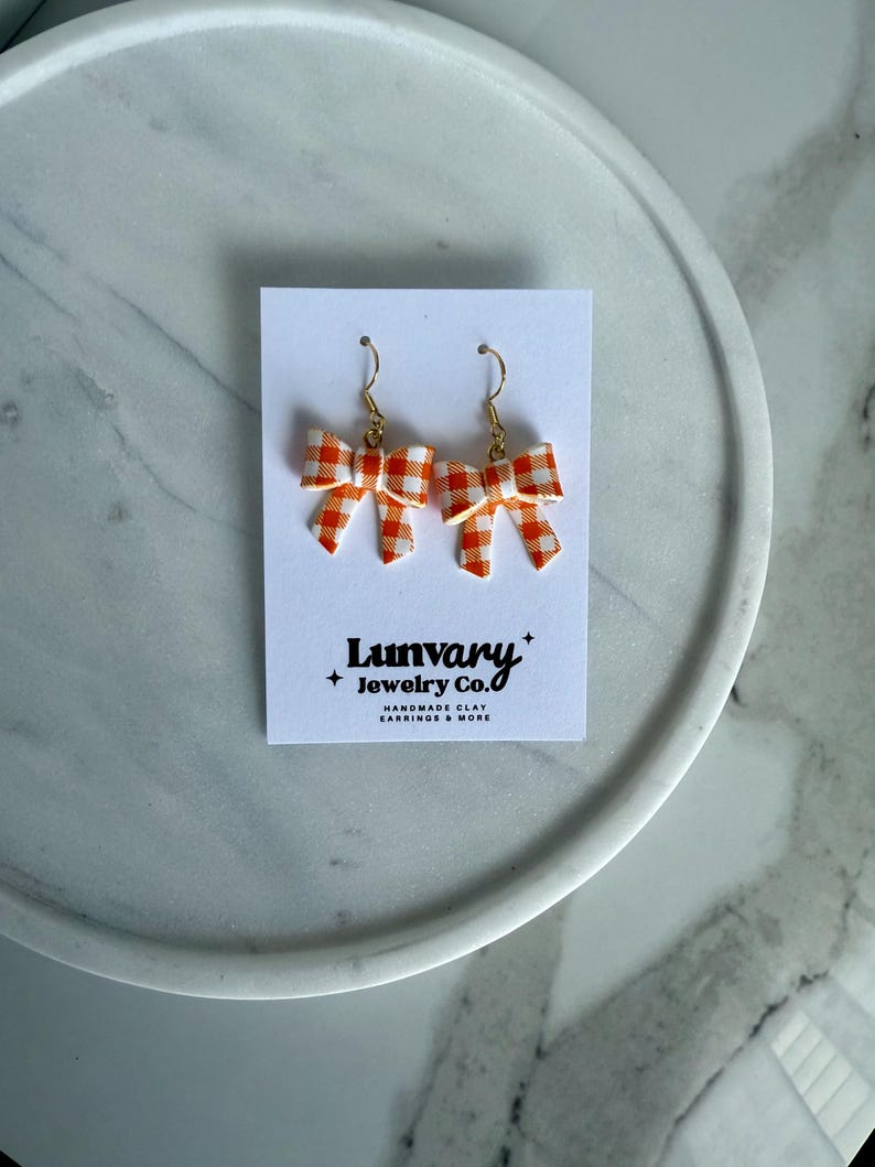 May include: A pair of bow-shaped earrings with a white and orange gingham pattern. The earrings are displayed on a white card with the text "Luvary Jewelry Co." and the words "HANDMADE CLAY EARRINGS & MORE". The earrings have gold-colored hooks.