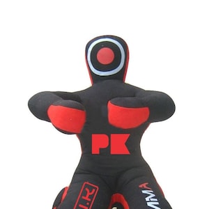 Puede incluir: Maniquí de entrenamiento negro y rojo para artes marciales. El maniquí tiene un objetivo en la cabeza y las letras "PK" en el torso. Las piernas tienen el texto "PARI.K" y "MMA".