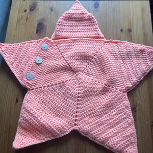 Puede incluir: Una manta para bebé en forma de estrella, de color coral, con capucha. La manta está hecha a mano y tejida a ganchillo con un punto texturizado. Cuenta con tres botones decorativos en la parte delantera. La capucha es puntiaguda.
