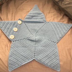 May include: A light blue star-shaped crochet blanket featuring a textured stitch pattern. Three large, round, white buttons are attached in a vertical row along one of the five points of the star, providing a decorative and functional closure detail.