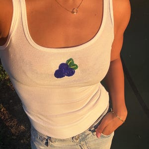 May include: A white ribbed tank top featuring a small, centered beaded patch of two blue berries with green leaves. The garment has a scoop neckline and is paired with light blue denim jeans. A thin gold chain necklace is worn around the neck.