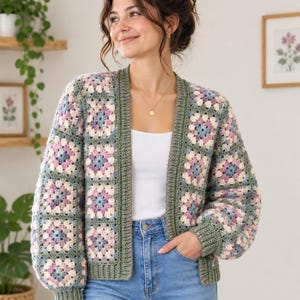 May include: A crochet cardigan constructed from multi-colored granny squares in shades of sage green, dusty pink, and periwinkle blue. The garment features a solid sage green ribbed trim along the front opening, cuffs, and hem, with long sleeves.