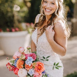 May include: A person in a white wedding dress smiles, holding a colourful bouquet. The bouquet includes orange, pink, and blue flowers with green leaves. The image includes the text "MACI COLLECTION" at the bottom.