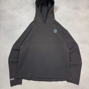 May include: Charcoal gray hoodie with a drawstring hood and a small blue and white emblem on the chest. The long-sleeved sweatshirt has a relaxed fit and a slightly cropped hemline. The sleeve cuff has a small white label.