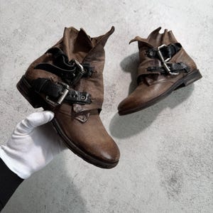 May include: Brown leather ankle boots with multiple black straps and silver buckles. The boots have a distressed look and a folded-over top. The boots are a neutral color and are suitable for various outfits.