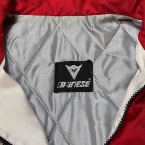 May include: Close-up of a red and white motorcycle jacket collar. The jacket has a quilted silver lining and a black label with the brand name "DAINESE". A black zipper runs down the front.