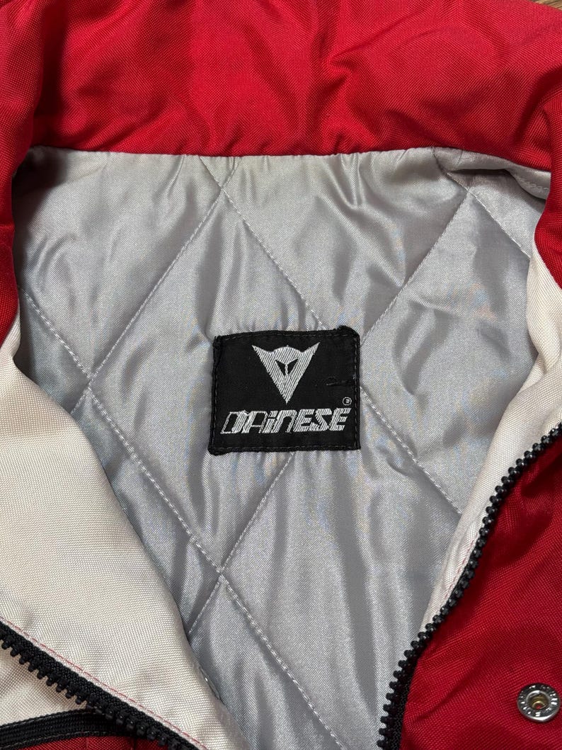 May include: Close-up of a red and white motorcycle jacket collar. The jacket has a quilted silver lining and a black label with the brand name "DAINESE". A black zipper runs down the front.