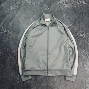 Puede incluir: Chaqueta deportiva gris claro con cremallera completa y cuello alzado. La chaqueta presenta rayas blancas en las mangas. La chaqueta está hecha de un material liso y brillante.