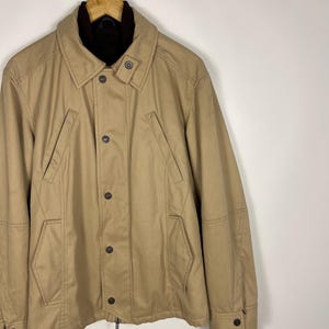 May include: A tan jacket with a dark brown collar and cuffs. The jacket has a button-down front, two front pockets, and a zipper closure. The jacket is hanging on a wooden hanger.