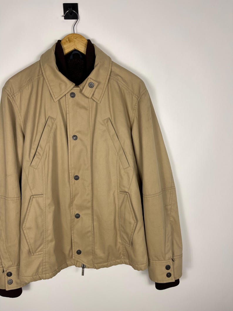 May include: A tan jacket with a dark brown collar and cuffs. The jacket has a button-down front, two front pockets, and a zipper closure. The jacket is hanging on a wooden hanger.