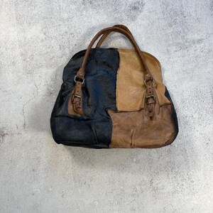May include: A patchwork leather tote bag in shades of black, tan, and brown. The bag features two brown leather handles and decorative buckles. The bag is designed with a unique color-block pattern.