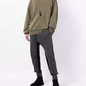 Puede incluir: Una sudadera verde oliva de gran tamaño con mangas largas y cuello redondo. El conjunto incluye pantalones grises oscuros con paneles negros y botines negros. Se ven un collar largo y un cuello de camisa a cuadros.