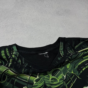Puede incluir: Camiseta negra con un dise&ntilde;o gr&aacute;fico verde y negro. El dise&ntilde;o es una ilustraci&oacute;n detallada de una criatura o monstruo. La camiseta est&aacute; hecha de un material suave y tiene cuello redondo.