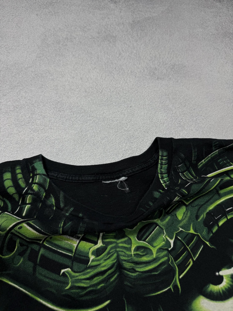 Puede incluir: Camiseta negra con un dise&ntilde;o gr&aacute;fico verde y negro. El dise&ntilde;o es una ilustraci&oacute;n detallada de una criatura o monstruo. La camiseta est&aacute; hecha de un material suave y tiene cuello redondo.