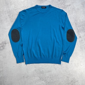 Puede incluir: Un jersey azul turquesa con mangas largas y cuello redondo. El jersey presenta coderas de color gris oscuro. La prenda se muestra sobre una superficie gris texturizada.