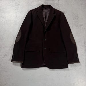 May include: A dark brown blazer with two front pockets, elbow patches, and buttoned cuffs. The jacket has a notched lapel and a three-button closure. The blazer is made of a soft, textured fabric.
