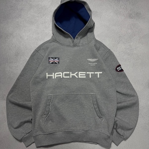 Puede incluir: Sudadera con capucha gris con capucha forrada en azul y bolsillo delantero. La sudadera con capucha presenta el texto "HACKETT" en blanco, un parche de la bandera británica y el logotipo de Aston Martin Racing. Un parche "53" está en la manga derecha.
