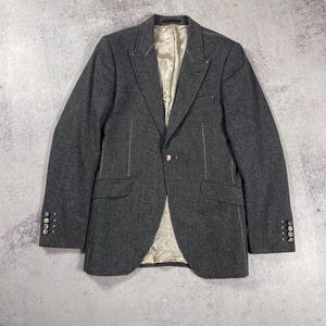 May include: A charcoal gray blazer with white stitching details and a single button closure. The blazer features a notched lapel, two front pockets, and decorative buttons on the sleeves. The interior lining is a light beige color.
