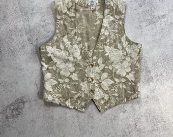 Puede incluir: Un chaleco beige con estampado floral, cuello en V y cierre de botones. El chaleco presenta un patrón de flores blancas y hojas verdes. El chaleco tiene un corte entallado y está hecho de una tela ligera.