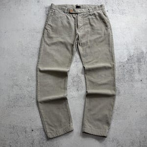 May include: A pair of light beige corduroy pants featuring a button-front closure, a waistband with belt loops, two side pockets, and a straight-leg cut. The fabric has a visible vertical ribbed texture throughout the garment.