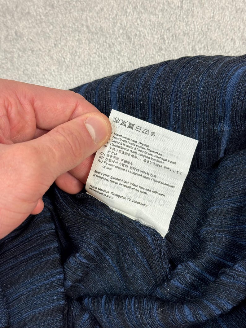 Puede incluir: Primer plano de una prenda de tejido acanalado azul marino con rayas azul oscuro. Se muestra una etiqueta blanca con instrucciones de cuidado, incluyendo lavado a mano y secado en plano.