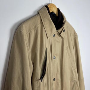 May include: A tan-colored jacket with a collared neckline and a dark brown interior lining. The jacket features a zipper pocket on the chest and button closures down the front. It is hanging on a wooden hanger.