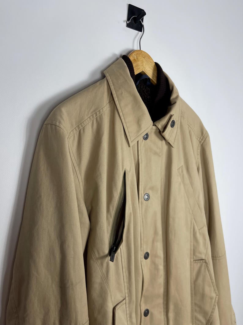 May include: A tan-colored jacket with a collared neckline and a dark brown interior lining. The jacket features a zipper pocket on the chest and button closures down the front. It is hanging on a wooden hanger.