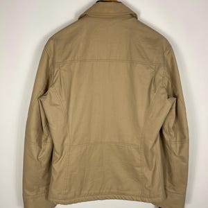 May include: Tan jacket with a collar and black cuffs, hanging on a wooden hanger. The jacket has a simple design with visible stitching and a structured fit. The back view showcases the jacket's tailored shape and neutral color.
