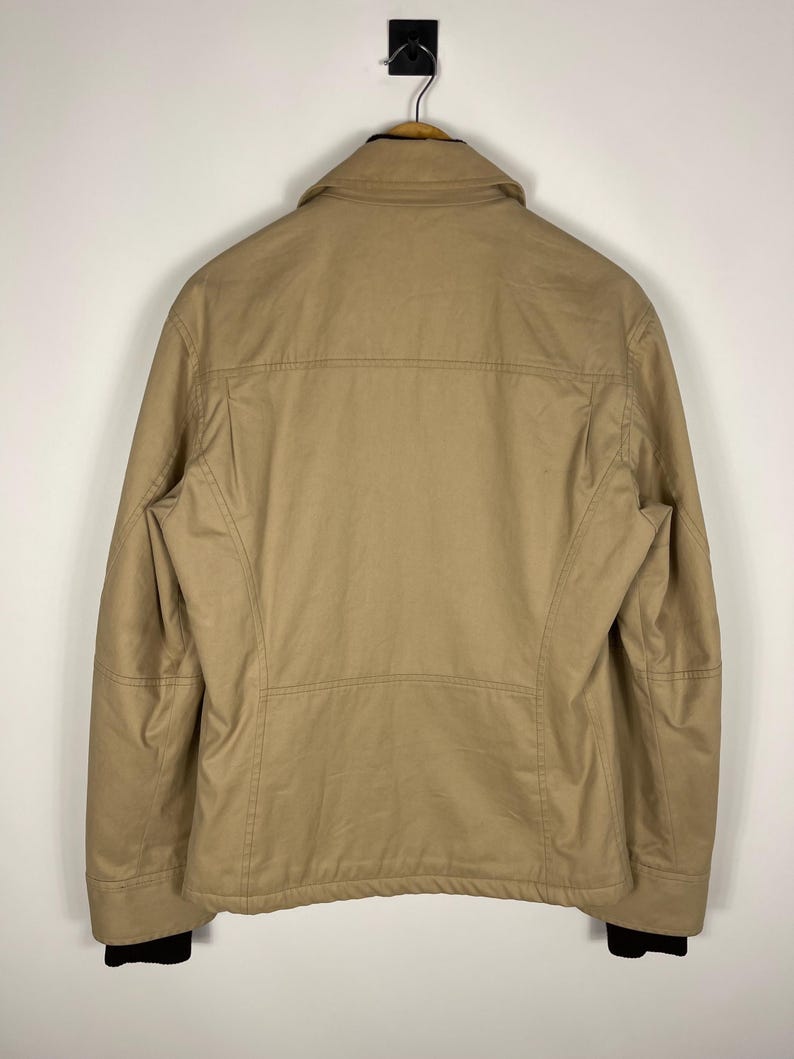 May include: Tan jacket with a collar and black cuffs, hanging on a wooden hanger. The jacket has a simple design with visible stitching and a structured fit. The back view showcases the jacket's tailored shape and neutral color.