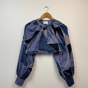 Puede incluir: Una blusa corta de manga larga con un diseño geométrico de patchwork en tonos azul marino, azul pizarra y gris claro. La prenda tiene cuello redondo, una tapeta de botones frontal y mangas fruncidas con puños abotonados.