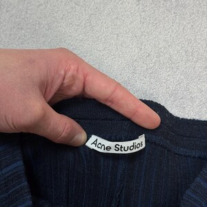 Puede incluir: Primer plano de una prenda de punto acanalado azul marino con la etiqueta blanca "Acne Studios" visible. La tela tiene rayas verticales. La prenda est&aacute; sujeta por una mano.
