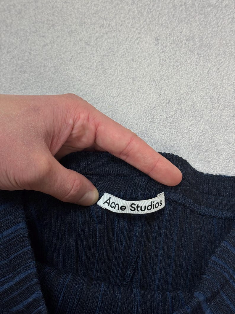 Puede incluir: Primer plano de una prenda de punto acanalado azul marino con la etiqueta blanca "Acne Studios" visible. La tela tiene rayas verticales. La prenda est&aacute; sujeta por una mano.