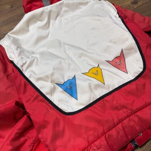 May include: Red and white jacket with a hood and a reflective stripe. The back features a white panel with three stylized arrow-shaped logos in blue, yellow, and red, outlined in black. The jacket has a red ribbed cuff.