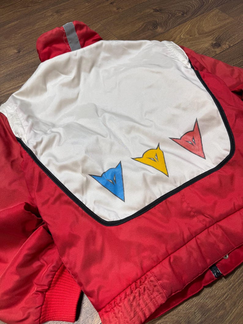May include: Red and white jacket with a hood and a reflective stripe. The back features a white panel with three stylized arrow-shaped logos in blue, yellow, and red, outlined in black. The jacket has a red ribbed cuff.