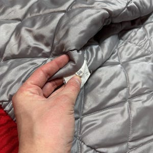 May include: Close-up of a quilted, silver-gray jacket lining. The lining has a diamond pattern and is made of a shiny material. A tag with the number 50 is visible. The jacket's outer layer is red.