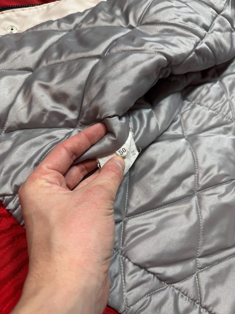 May include: Close-up of a quilted, silver-gray jacket lining. The lining has a diamond pattern and is made of a shiny material. A tag with the number 50 is visible. The jacket's outer layer is red.
