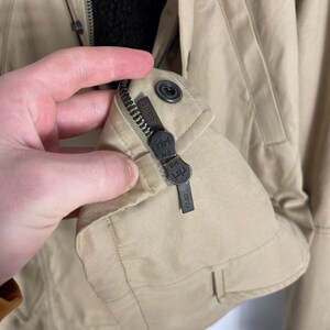 May include: Tan jacket with a black interior lining. The jacket has a zipper closure and a small metal tag with the brand name. The jacket is made of a durable material and has a classic design.