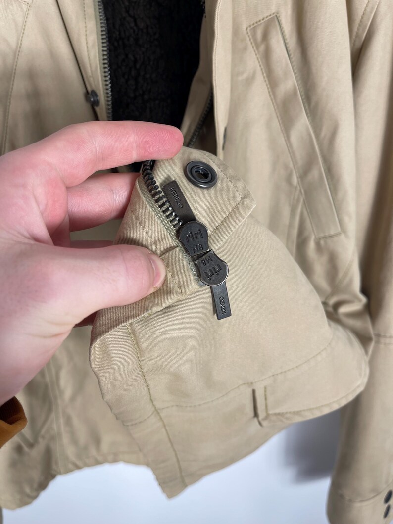 May include: Tan jacket with a black interior lining. The jacket has a zipper closure and a small metal tag with the brand name. The jacket is made of a durable material and has a classic design.