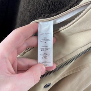 May include: Close-up of a beige jacket with a brown lining. The jacket has a zipper and a button closure. A tag with text in multiple languages is visible, including "100% Coton-Cotton" and "69% Acrylique-Acrylic".