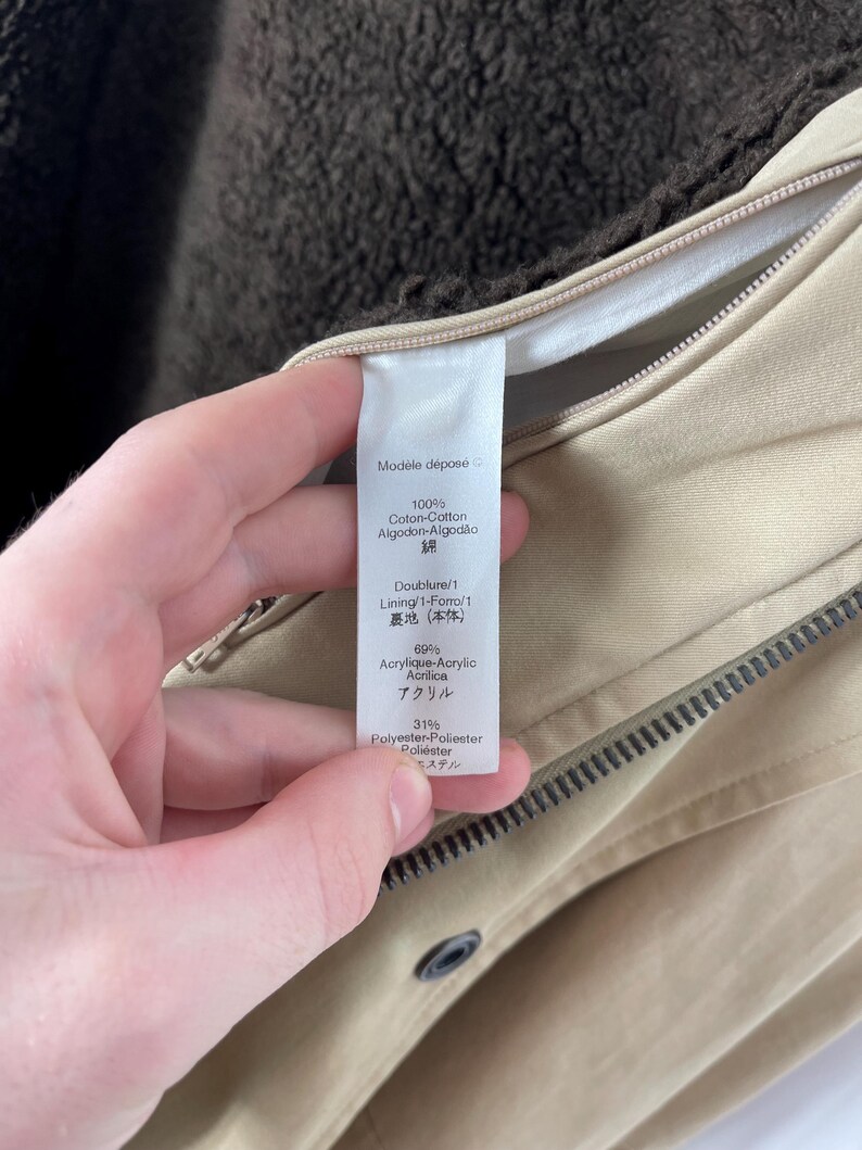 May include: Close-up of a beige jacket with a brown lining. The jacket has a zipper and a button closure. A tag with text in multiple languages is visible, including "100% Coton-Cotton" and "69% Acrylique-Acrylic".